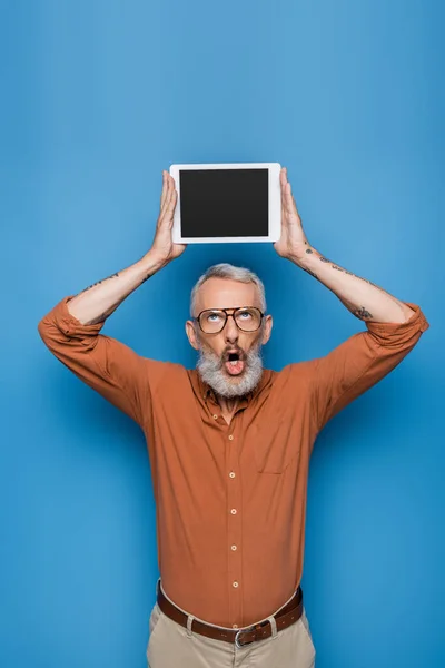 Sorprendido hombre de mediana edad en gafas y camisa celebración tableta digital con pantalla en blanco por encima de la cabeza en azul - foto de stock