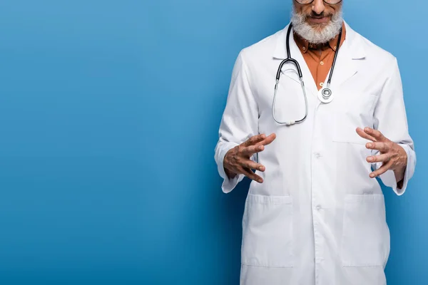 Cropped view of bearded middle aged doctor in white coat gesturing on blue — Stock Photo