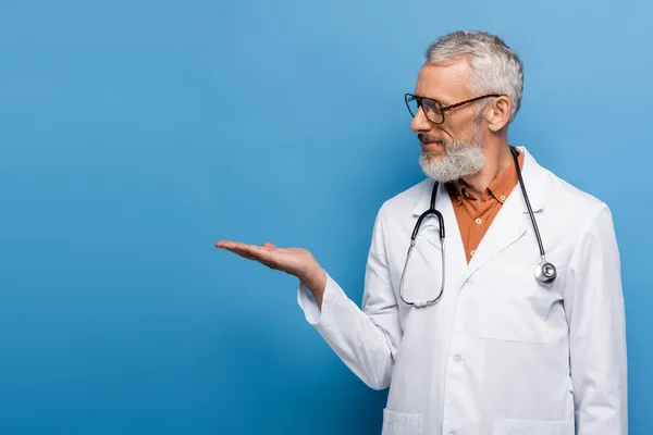 Bärtiger Arzt mittleren Alters mit Brille und weißem Mantel zeigt mit der Hand auf blau — Stockfoto