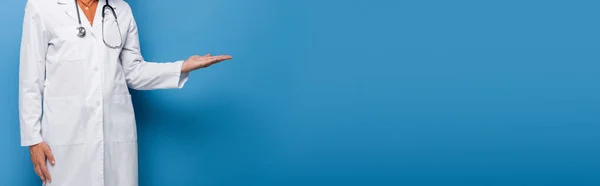 Vista recortada del médico en bata blanca apuntando con la mano en azul, bandera - foto de stock