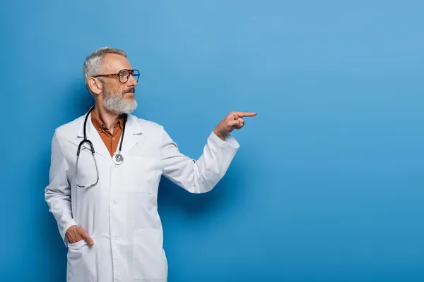 Barbu médecin d'âge moyen en manteau blanc pointant avec le doigt et debout avec la main dans la poche sur bleu — Photo de stock