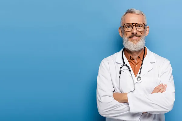 Allegro medico di mezza età in cappotto bianco in piedi con braccia incrociate su blu — Foto stock