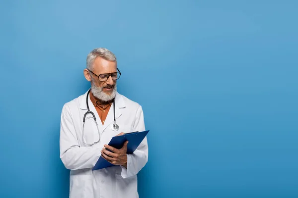 Médecin d'âge moyen en manteau blanc et lunettes écriture prescription sur presse-papiers sur bleu — Photo de stock