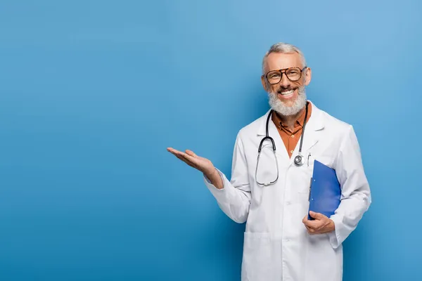 Happy middle aged doctor in white coat and glasses holding clipboard and pointing with hand on blue — Stock Photo