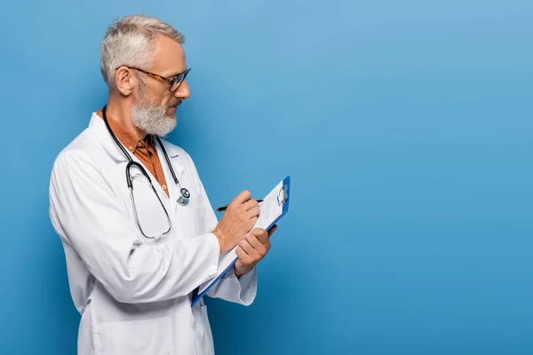 Barbudo médico de meia idade em casaco branco e óculos de prescrição escrita na área de transferência em azul — Fotografia de Stock