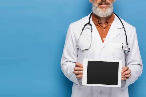 Ausgeschnittene Ansicht eines bärtigen Arztes mittleren Alters im weißen Mantel, der ein digitales Tablet mit leerem Bildschirm auf blauem Hintergrund hält — Stockfoto