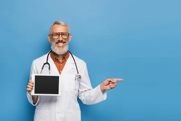 Happy middle aged doctor in white coat holding digital tablet with blank screen while pointing with finger on blue — Stock Photo