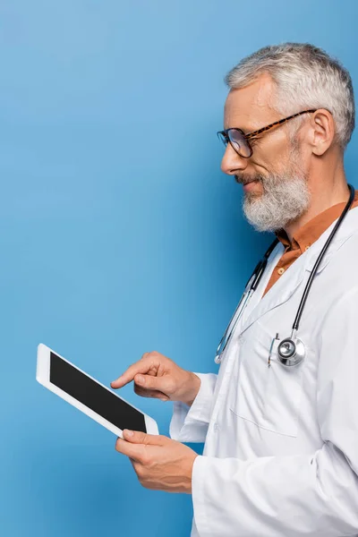 Smiling middle aged doctor in white coat pointing at digital tablet with blank screen on blue — Stock Photo