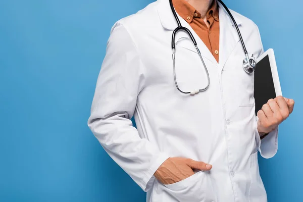 Vue recadrée du médecin en manteau blanc debout avec la main dans la poche et tenant tablette numérique avec écran blanc sur bleu — Photo de stock