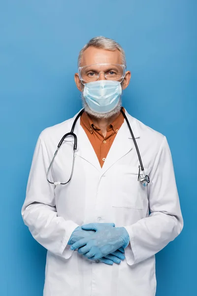 Middle aged doctor in white coat, medical mask and goggles standing on blue — Stock Photo