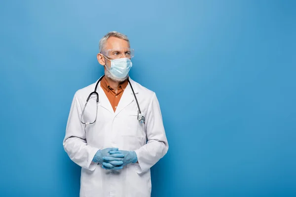 Middle aged doctor in white coat, medical mask and goggles standing with clenched hands on blue — Stock Photo