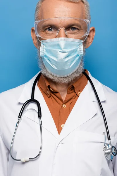 Middle aged doctor in white coat, medical mask and goggles isolated on blue — Stock Photo