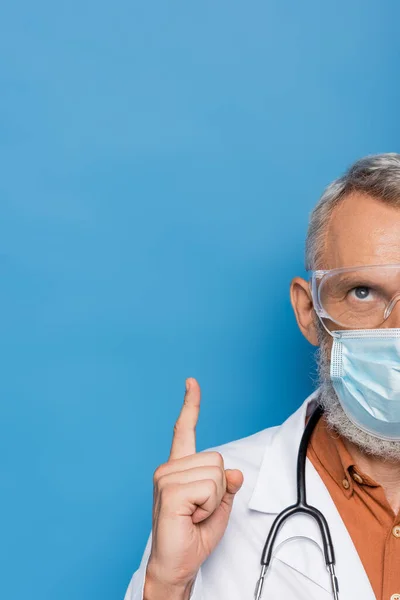 Cropped view of middle aged doctor in medical mask and goggles pointing up on blue — Stock Photo