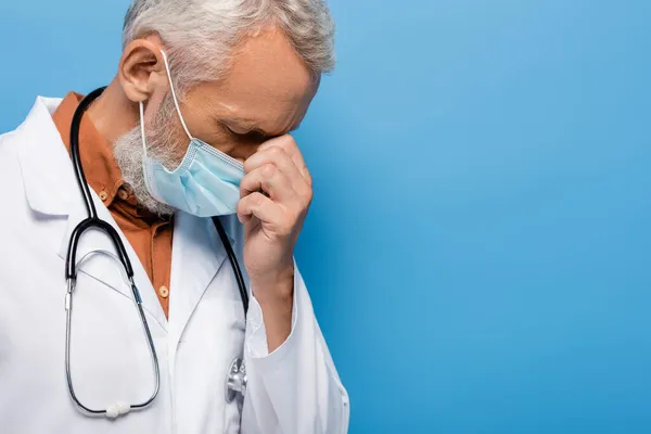 Médico de mediana edad trastornado en bata blanca y máscara médica en azul - foto de stock