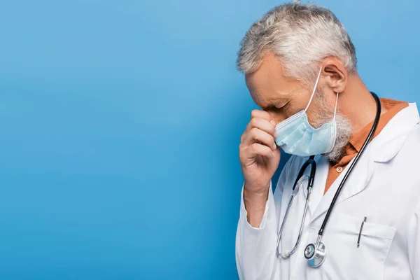 Angespannter Arzt mittleren Alters in weißem Mantel und medizinischer Maske auf blau — Stockfoto