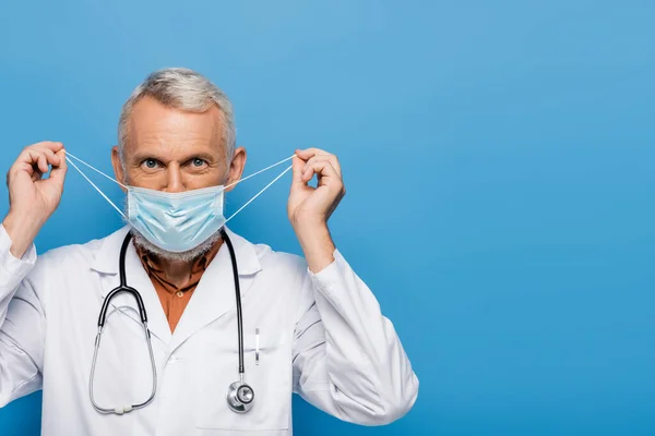Médico de meia idade em casaco branco ajustando máscara médica isolada em azul — Fotografia de Stock
