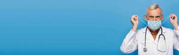 Medico di mezza età in cappotto bianco regolazione maschera medica isolata su blu, banner — Foto stock