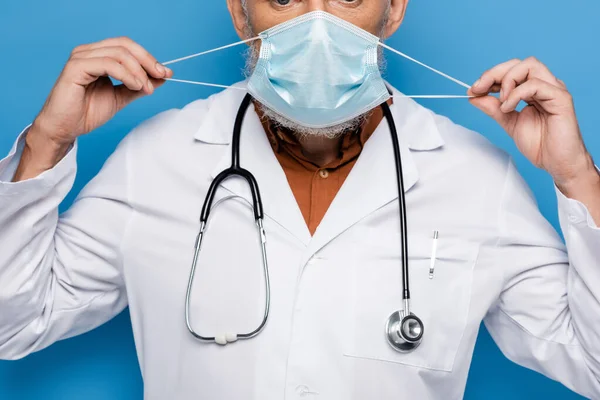 Ausgeschnittene Ansicht eines Arztes mittleren Alters in weißem Mantel, der die medizinische Maske auf blau isoliert anpasst — Stockfoto