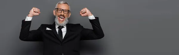 Hombre de mediana edad excitado en traje y gafas regocijándose en gris, bandera - foto de stock