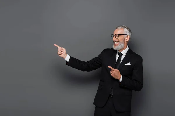 Alegre hombre de negocios de mediana edad en traje y gafas apuntando con los dedos en gris - foto de stock