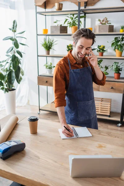 Lächelnder Verkäufer spricht auf Smartphone und schreibt auf Notizbuch im Blumenladen — Stockfoto