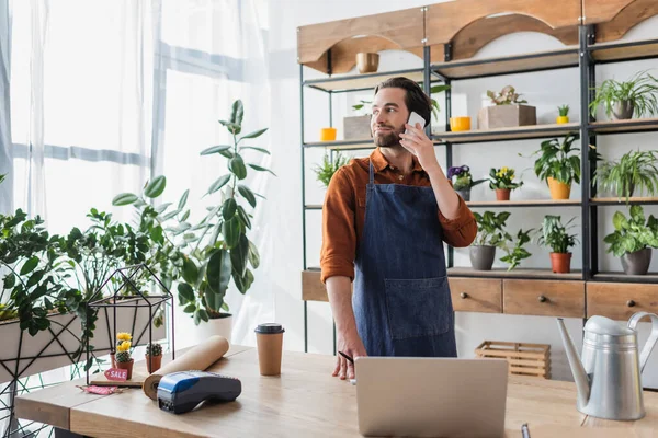 Venditore in grembiule parlando su smartphone vicino a caffè e laptop in negozio di fiori — Foto stock