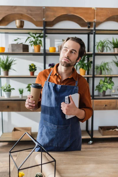 Vendedor en delantal sosteniendo taza de papel y cuaderno en tienda de flores - foto de stock