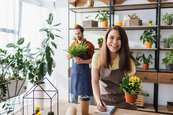Cheerful seller in apron looking at camera while holding plant and writing on notebook in flower shop — Stock Photo