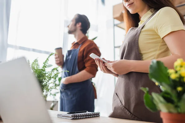 Seller in apron using smartphone near notebook and laptop in flower shop — Stock Photo