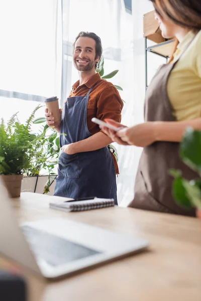 Rivenditore sorridente con bicchiere di carta guardando collega con smartphone nel negozio di fiori — Foto stock