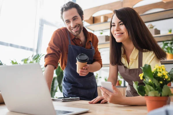 Positive sellers in aprons holding smartphone and paper cup in flower shop — Stock Photo