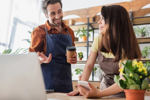 Smiling seller holding smartphone and looking at colleague with coffee to go near laptop in flower shop — Stock Photo