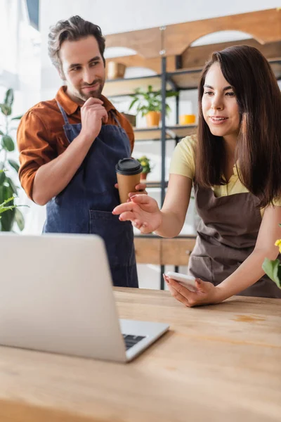 Seller in apron holding smartphone and pointing at laptop near colleague with paper cup in flower shop — Stock Photo