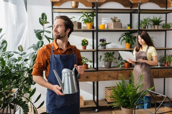 Junger Florist hält Gießkanne in der Nähe von Pflanzen und verschwommener Kollege im Blumenladen — Stockfoto