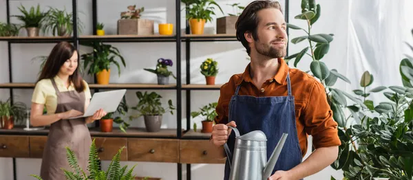 Vendedor celebración de regadera cerca de las plantas y colega borrosa en la tienda, pancarta - foto de stock