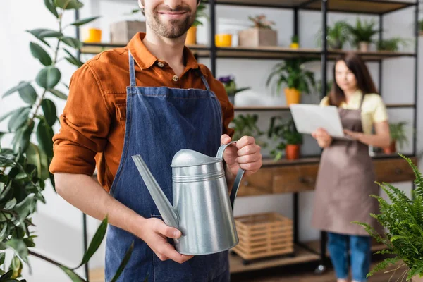 Smiling seller holding watering can near plants and colleague in flower shop — Stock Photo
