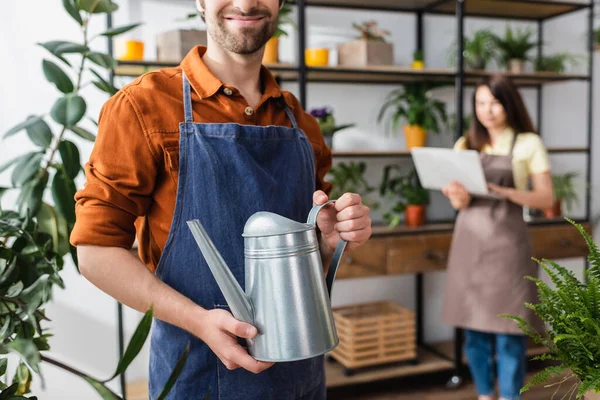 Vendedor sonriente sosteniendo regadera cerca de plantas y colega en floristería - foto de stock