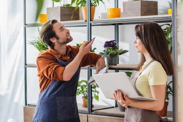 Floristería sosteniendo portátil cerca de colega apuntando a las plantas en los estantes en la tienda - foto de stock