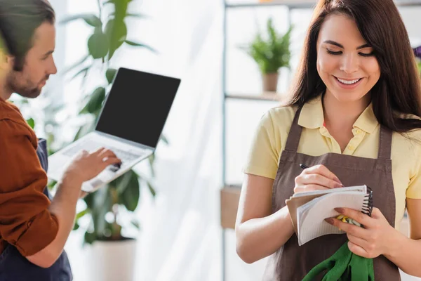 Alegre florista escribiendo en un cuaderno cerca de un colega borroso con computadora portátil en la tienda de flores - foto de stock