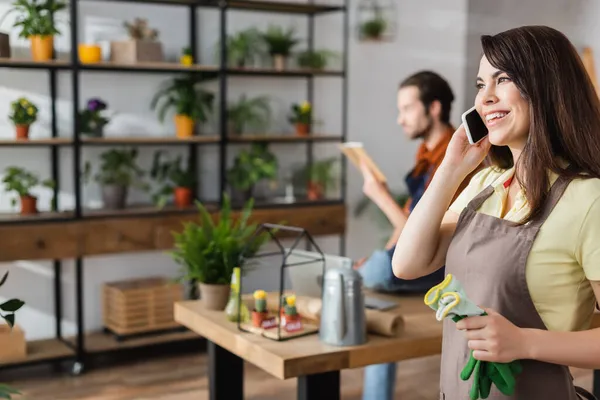 Venditore sorridente che tiene guanti da giardinaggio e parla su smartphone in negozio di fiori — Foto stock