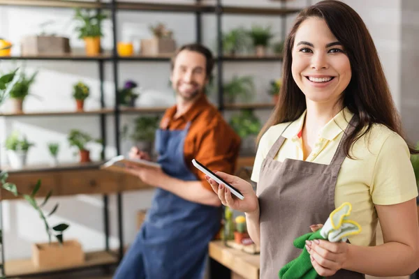 Positive florist holding garden gloves and smartphone in flower shop — Stock Photo