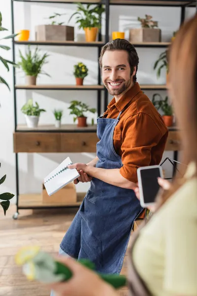 Fleuriste souriant tenant un carnet et regardant un collègue flou avec smartphone dans un magasin de fleurs — Photo de stock