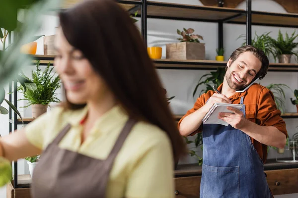 Alegre florista hablando en el teléfono inteligente y escribiendo en el cuaderno cerca de colega borroso en la tienda - foto de stock