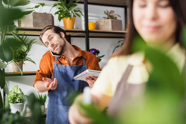 Florist holding notebook and talking on smartphone near blurred colleague in flower shop — Stock Photo