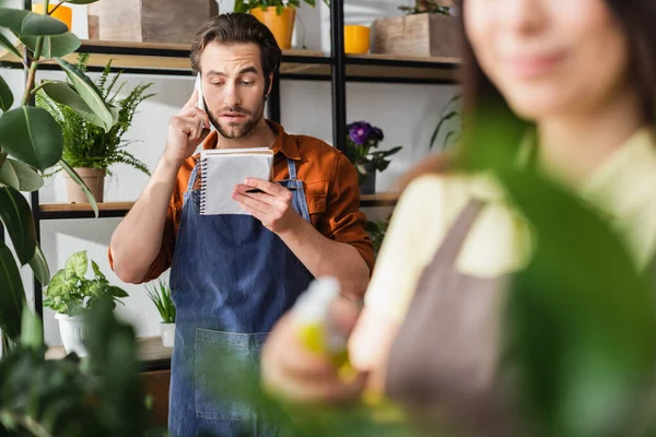 Joven florista hablando por teléfono celular y mirando el cuaderno cerca de las plantas y colega borrosa en la tienda - foto de stock