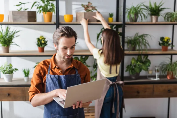 Floristin mit Laptop neben verschwommenem Kollegen mit Pflanzen im Geschäft — Stockfoto