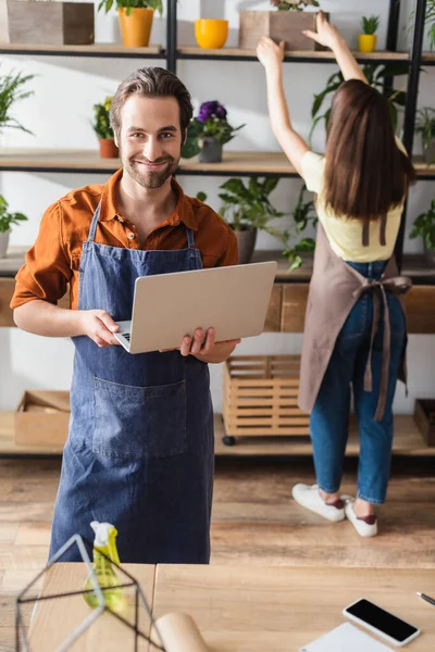 Fiorista sorridente che tiene il computer portatile e guarda la macchina fotografica vicino a collega in negozio di fiori — Foto stock