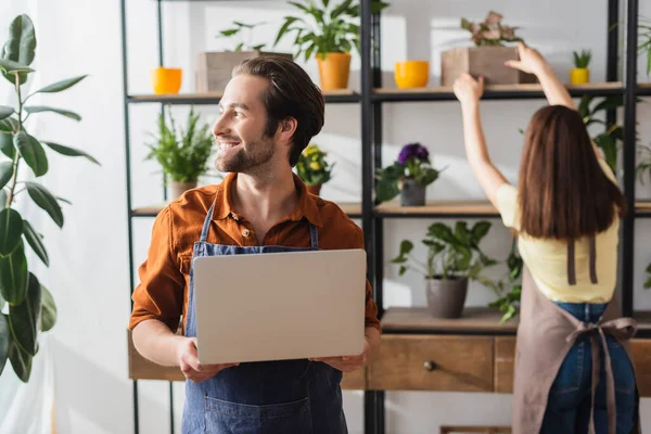 Fröhliche Verkäuferin in Schürze hält Laptop neben Kollegin mit Pflanzen im Blumenladen — Stockfoto