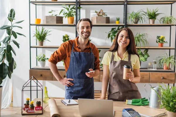 Fioristi sorridenti con caffè e smartphone guardando la fotocamera vicino alle piante nel negozio — Foto stock