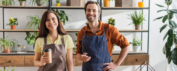 Cheerful florists in aprons holding coffee to go and smartphone in flower shop, banner — Stock Photo